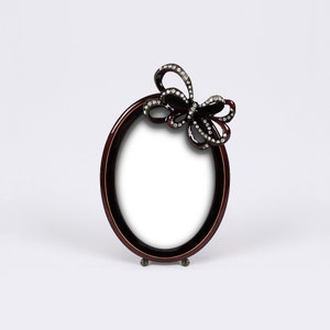 May include: An oval-shaped picture frame with a dark red finish and a decorative jeweled bow. The frame has a gold trim and stands on a small base.