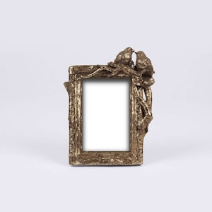 May include: Gold-toned picture frame with a decorative design of a tree branch with two birds perched on it. The frame has a rectangular opening for a photo.