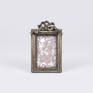 May include: A silver picture frame with a decorative bow design at the top. The frame has a light pink floral pattern inside.