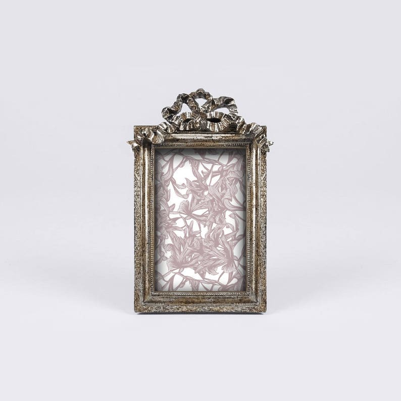 May include: A silver picture frame with a decorative bow design at the top. The frame has a light pink floral pattern inside.