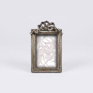 May include: A silver-toned picture frame with an ornate bow design at the top. The frame has a rectangular opening for a photo and a floral pattern inside the frame.