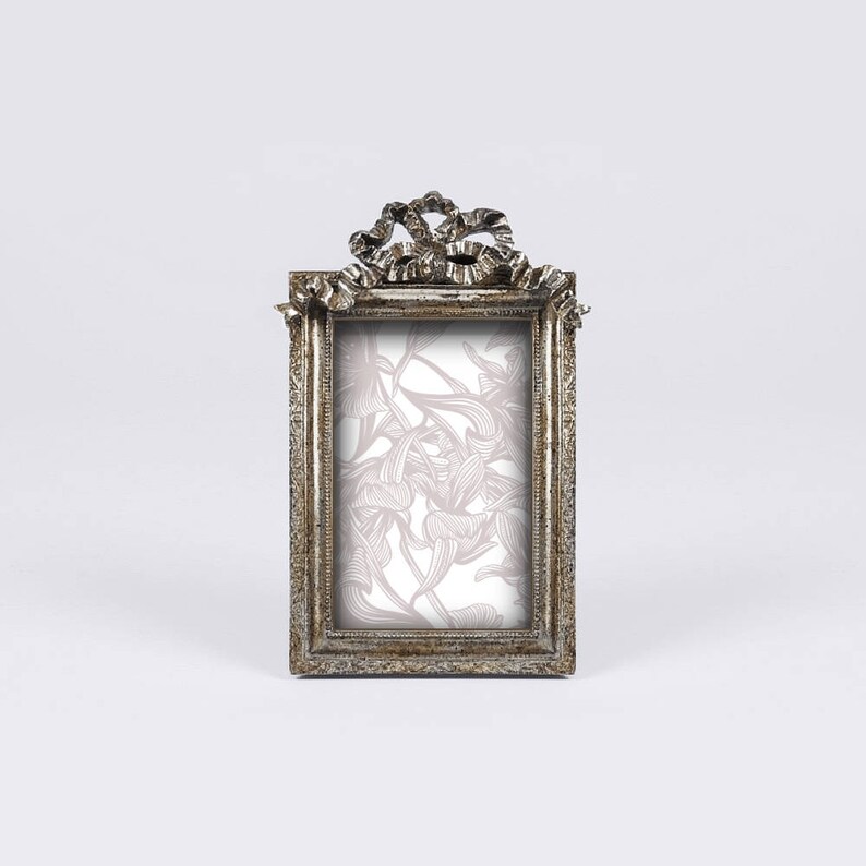 May include: A silver-toned picture frame with an ornate bow design at the top. The frame has a rectangular opening for a photo and a floral pattern inside the frame.