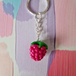 May include: A silver keyring with a miniature raspberry charm. The raspberry is a bright pink with a green leafy top. The keyring has a silver ring and chain. The background features colourful brushstrokes of pink, purple, and blue.