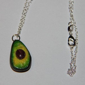 Peut inclure: Un collier en chaîne argent avec un pendentif avocat vert et jaune. L'avocat est coupé en deux et a un noyau brun à l'intérieur.