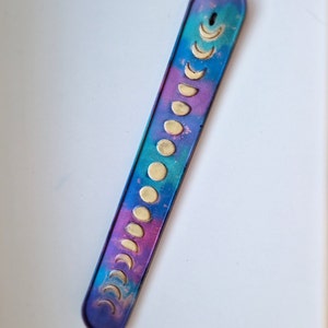 May include: A purple, blue, and pink incense holder with a galaxy-like design. The holder has a row of gold crescent moons along the length of the holder.