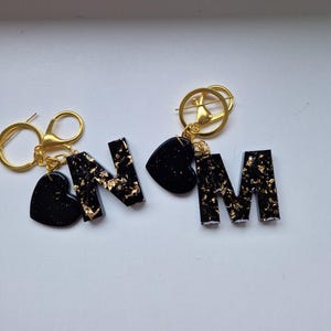 Könnte beinhalten: Zwei Schlüsselanhänger mit goldfarbener Hardware. Einer zeigt den Buchstaben "N" und einen herzförmigen Anhänger, beide schwarz mit Goldflocken. Der andere Schlüsselanhänger hat den Buchstaben "M" und einen Herzanhänger, ebenfalls schwarz mit Goldflocken.