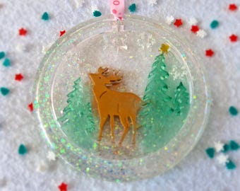 May include: A clear round Christmas ornament with a gold glitter background. Inside the ornament is a brown reindeer standing in a snowy forest with green trees. The ornament has a white ribbon with the word "HO" printed on it.