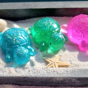 May include: Four small, glittery turtle figurines in a variety of colours, including blue, green, and pink. The turtles are arranged on a white sandy surface with seashells and a starfish.