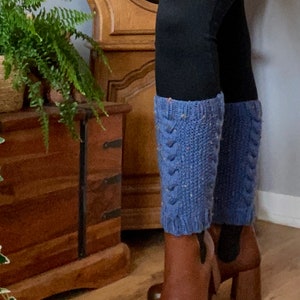 Puede incluir: Un par de polainas de punto azul con un patrón de punto de cable. Las polainas se llevan sobre unos leggings negros y unas botas de tacón marrón.