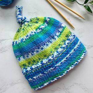 May include: A hand-knitted baby hat in a striped pattern of blue, green, and white yarn. The hat has a knotted top and a soft, textured feel.