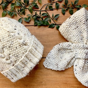 Puede incluir: Un gorro blanco de punto con un pompón esponjoso y una bufanda blanca de punto a juego. Ambos artículos tienen un patrón moteado.