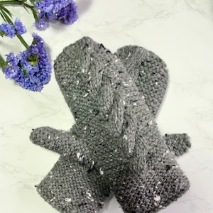 May include: A pair of gray knitted mittens with a white fleck pattern. The mittens are folded together and have a cable knit design.