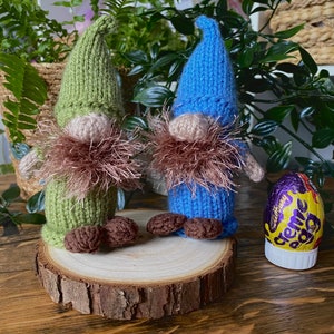 Puede incluir: Dos adornos de gnomo tejidos, uno verde y otro azul, con barbas de lana marrón. Los gnomos están sentados sobre una superficie de madera con un huevo de chocolate en el fondo.