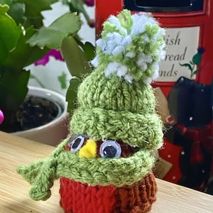 May include: A knitted robin wearing a green hat with a white pom-pom and a green scarf. The robin has large googly eyes and a yellow beak.