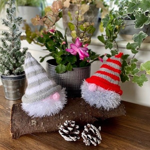 May include: Two knitted gnomes with white fuzzy beards. One gnome has a red and white striped hat, the other has a gray and white striped hat. The gnomes are sitting on a wooden log with pine cones on the table.