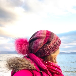 Può includere: Una donna indossa un berretto a maglia a righe rosa e marrone con un pompon rosa soffice. Il berretto è a maglia e ha una vestibilità morbida. La donna è in piedi di fronte a uno specchio d'acqua e un cielo nuvoloso.