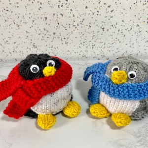 Puede incluir: Dos adornos de pingüino tejidos con bufandas. Un pingüino tiene una bufanda roja y el otro una bufanda azul. Ambos pingüinos tienen ojos negros y picos amarillos.