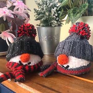 May include: Two knitted snowman ornaments with red and black striped scarves and gray and black knit hats with red pom poms.