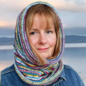 May include: A woman wearing a colorful striped knitted cowl neck scarf. The scarf is made of a soft, textured yarn and has a variety of colors, including blue, green, yellow, and red. The woman is wearing a blue denim jacket.