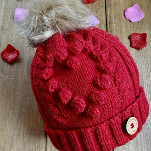 Könnte beinhalten: Rote Strickmütze mit Bommel und einem Knopf aus Holz. Die Mütze hat ein Herzmuster und ist aus dickem Garn gefertigt.