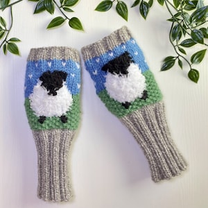 Pode incluir: Um par de luvas sem dedos de malha cinzenta com um design de ovelha azul, verde e branca. A ovelha tem a cara preta e lã branca e fofa.
