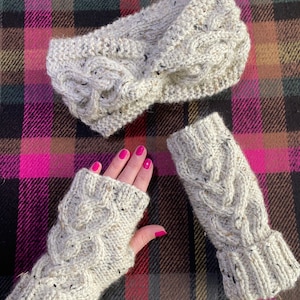 May include: A cream-coloured knitted headband and fingerless gloves set. The cable-knit design features flecks of dark thread. The gloves are shown on a hand with pink nail polish. The set is displayed on a plaid background.