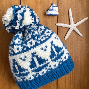 May include: A blue and white knitted hat with a white pom-pom. The hat has a pattern of sailboats.