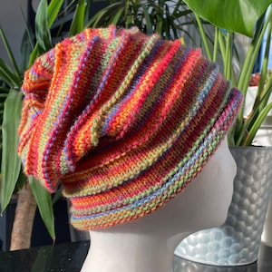 May include: A colourful striped knitted beanie hat with a slouchy fit. The hat is made with a variety of colours, including red, orange, yellow, green, and blue.