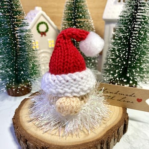May include: A knitted Santa Claus ornament with a red hat, white beard, and a fluffy white pom-pom on top. The ornament is sitting on a wooden slice with a tag that says "Handmade with Love".