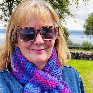 Puede incluir: Una mujer con una chaqueta vaquera y una bufanda de punto a rayas de color morado, rosa y azul. Lleva gafas de sol de carey y tiene el pelo rubio.