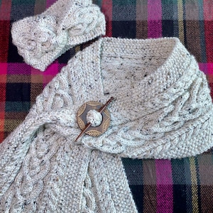 May include: A white knitted scarf and headband set with a cable knit pattern and a wooden pin with a decorative circle. The set is displayed on a plaid fabric background.