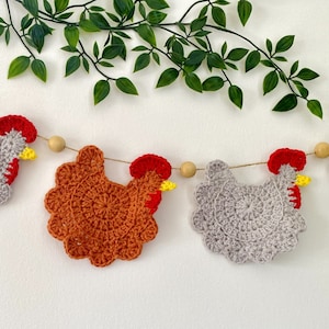 Peut inclure: Une guirlande de poulets en crochet avec des poulets gris et orange alternés. Les poulets ont des crêtes rouges et des becs jaunes. La guirlande est enfilée sur une ficelle et des perles en bois.