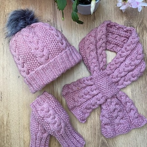 Könnte beinhalten: Eine rosa Strickmütze mit flauschigem Bommel, passende fingerlose Handschuhe und ein Zopfmuster-Schal. Das Set ist aus weichem Garn gefertigt und hat eine zarte rosa Farbe.