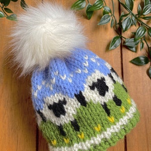 Könnte beinhalten: Eine gestrickte Mütze mit einem weißen Kunstfell-Bommel. Die Mütze hat ein blaues Himmelsdesign mit weißen Schneeflockenmustern, grünem Gras und schwarz-weißen Schafen. Die Mütze ist aus weichem Garn gefertigt.