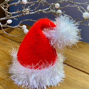 May include: A red knitted Santa hat with a white fluffy trim. The hat is sitting on a wooden surface.
