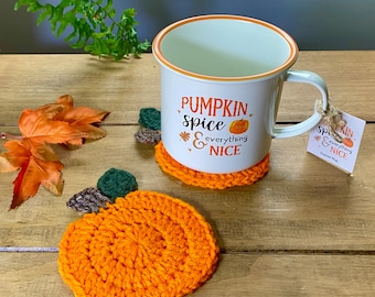 May include: Two crocheted pumpkin-shaped coasters in orange with green and brown leaves. A white enamel mug with the text "Pumpkin Spice & everything nice" is sitting on one of the coasters.