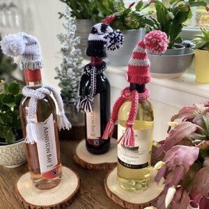 Può includere: Tre bottiglie di vino con cappelli e sciarpe di lana. Le bottiglie sono su sottobicchieri di legno. Le bottiglie sono etichettate "Arrington Bay Rose", "Aston Bay" e "Chardonnay".
