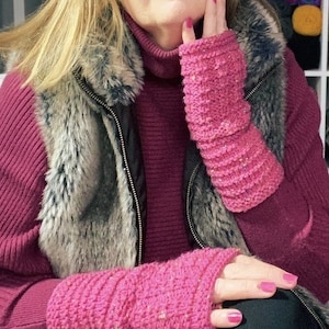 May include: A pair of pink knitted fingerless gloves with a textured pattern. The gloves are worn by a person with a pink sweater and a faux fur vest.