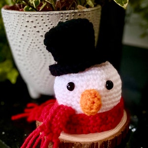 Peut inclure: Un bonhomme de neige en crochet portant un chapeau haut de forme noir et une écharpe rouge. Le bonhomme de neige est assis sur une tranche de bois.