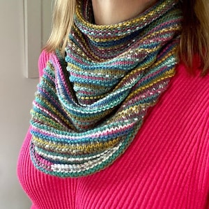 May include: A multi-colored knitted cowl scarf with horizontal stripes in shades of pink, blue, green, yellow, and white. The scarf is draped around the neck and shoulders, showcasing the textured knit design. The background is a solid pink top.