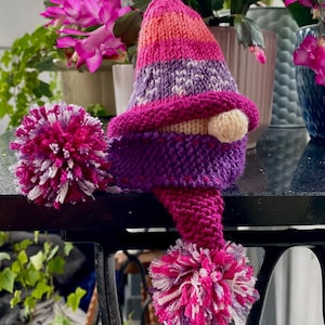 Pode incluir: Um gnomo de malha rosa e roxo com um pompom branco na ponta do chapéu e um pompom branco na ponta do cachecol.