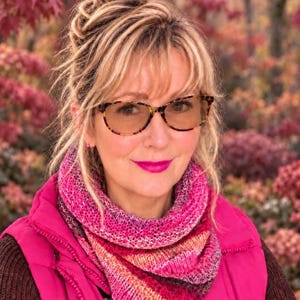 May include: A hand-knitted cowl scarf featuring a variegated colour palette of magenta, pink, orange, and deep purple. The accessory is worn over a dark brown jumper and a bright magenta vest, showcasing a textured knit pattern and soft yarn.