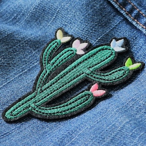 Könnte beinhalten: Ein gestickter Aufnäher mit einem grünen Kaktus mit rosa und grünen Blüten. Der Aufnäher ist auf eine blaue Jeansjacke genäht.