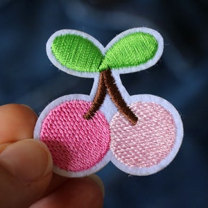 Puede incluir: Un parche de fieltro blanco con un diseño bordado de dos cerezas rosas con hojas verdes y un tallo marrón.
