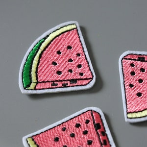 May include: Three embroidered watermelon slice patches. The patches are pink with black seeds and green rind. The patches are on a grey background.