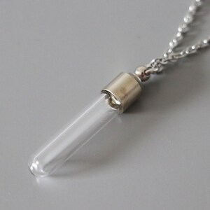 May include: A clear glass vial pendant necklace with a silver-colored metal cap and chain. The vial is cylindrical and empty, suspended from a delicate chain. The background is a solid gray.