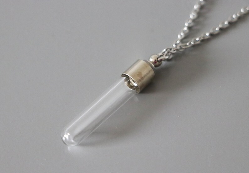 May include: A clear glass vial pendant necklace with a silver-colored metal cap and chain. The vial is cylindrical and empty, suspended from a delicate chain. The background is a solid gray.