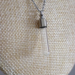 May include: A clear glass vial pendant necklace with a silver-colored metal cap and chain. The vial is cylindrical and hangs vertically. The necklace is displayed on a beige textured surface.