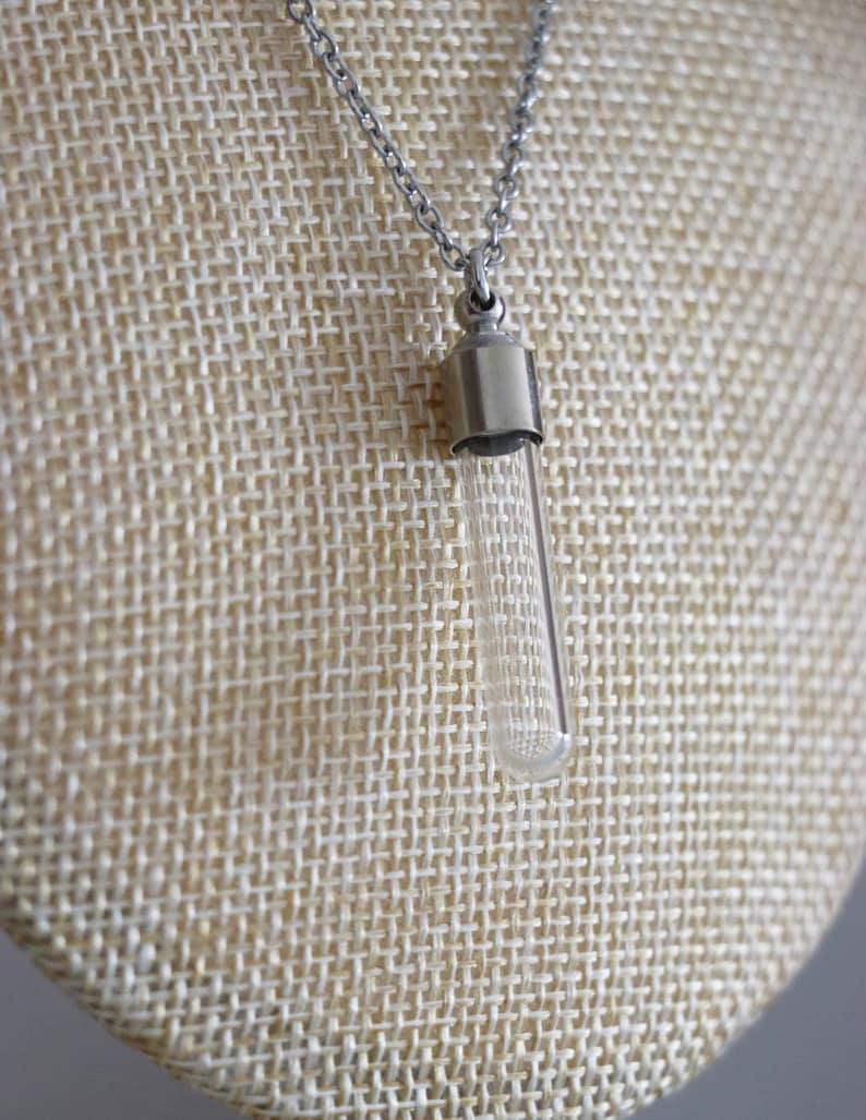 May include: A clear glass vial pendant necklace with a silver-colored metal cap and chain. The vial is cylindrical and hangs vertically. The necklace is displayed on a beige textured surface.