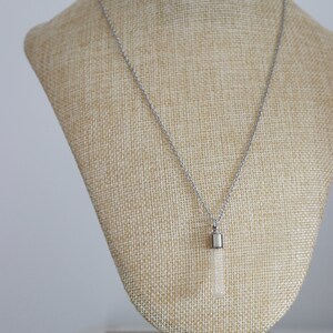 May include: A silver necklace with a rectangular, clear pendant. The pendant is suspended from a delicate chain and is displayed on a beige bust. The necklace is a simple, elegant piece of jewelry.